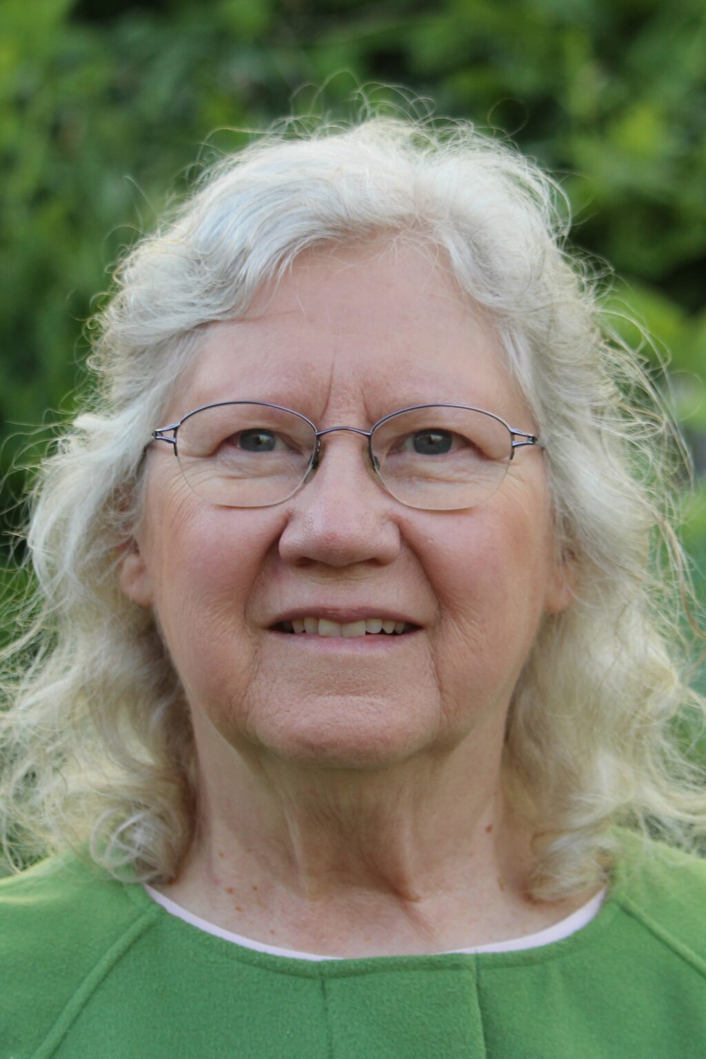 Smiling elderly woman with glasses and curly white hair outdoors.